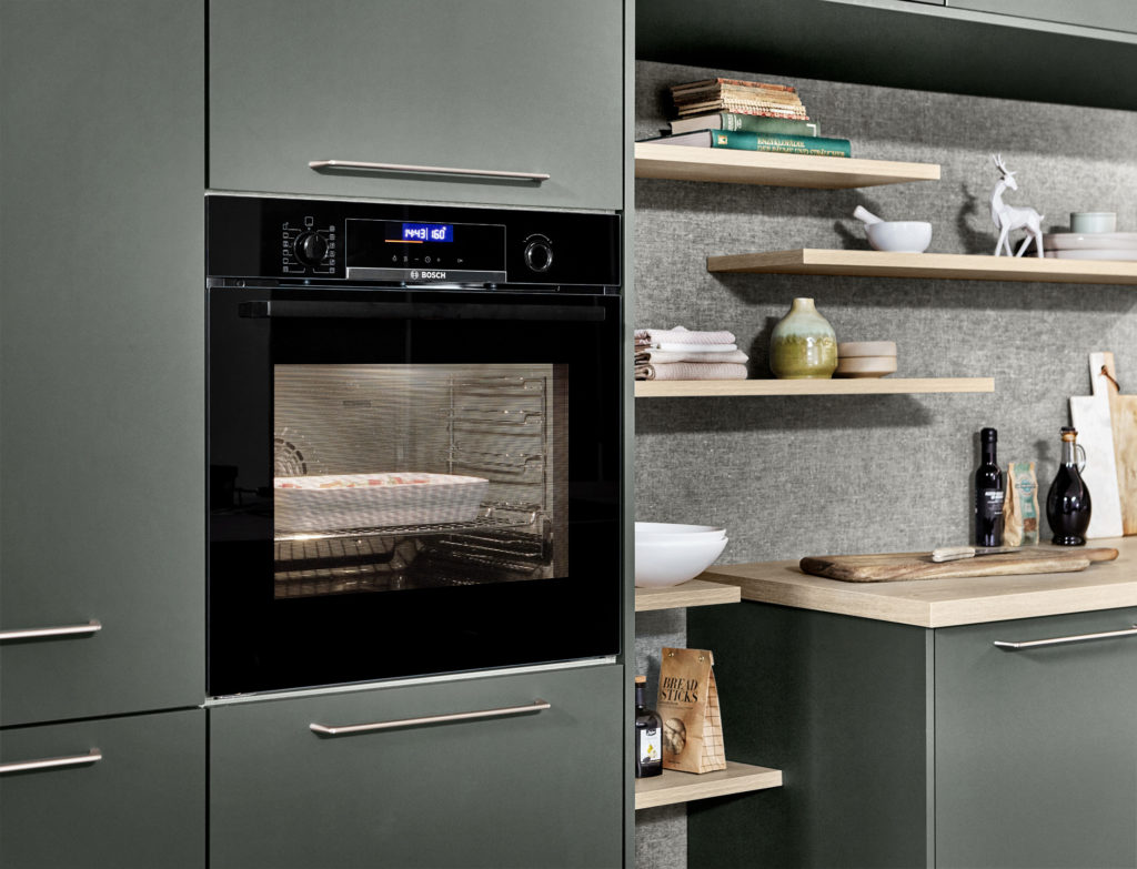 Horno encendido en cocina de acabado verde bosque y tirador largo de acero inoxidable. Cocina con encimera de madera.